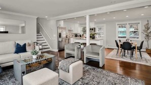 A modern living room with a sofa, ottoman, coffee table, and chairs. There is a staircase leading to a kitchen and dining area. - 5BR Beach House, Water Views, Game Room, Backyard (Stamford)