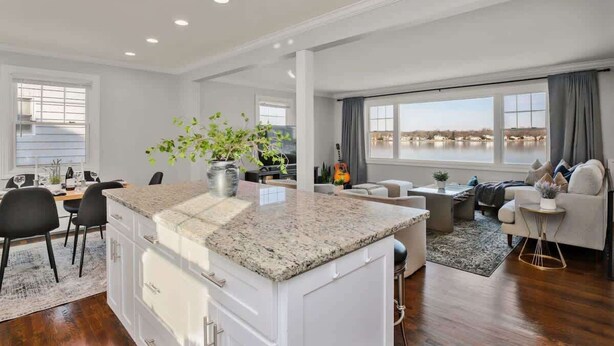 A modern kitchen with granite countertops, a dining area with black chairs, a living room with a sofa and a coffee table, and a large window with a view. - 5BR Beach House, Water Views, Game Room, Backyard (Stamford)