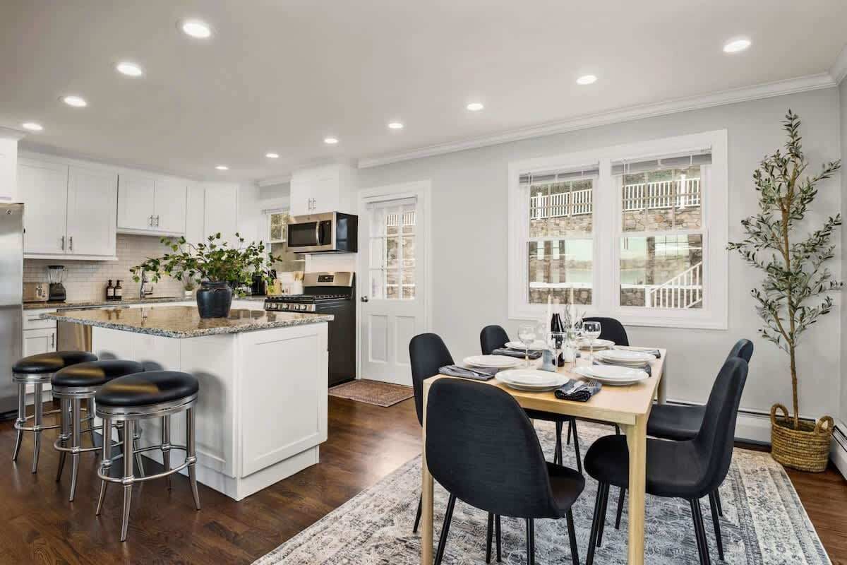 A modern kitchen with white cabinets, a central island, bar stools, and a dining area with black chairs.