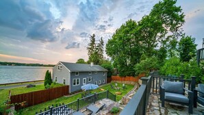 A residential backyard with a stone patio, outdoor furniture, a wooden fence, and a view of a lake. - 5BR Beach House, Water Views, Game Room, Backyard (Stamford)