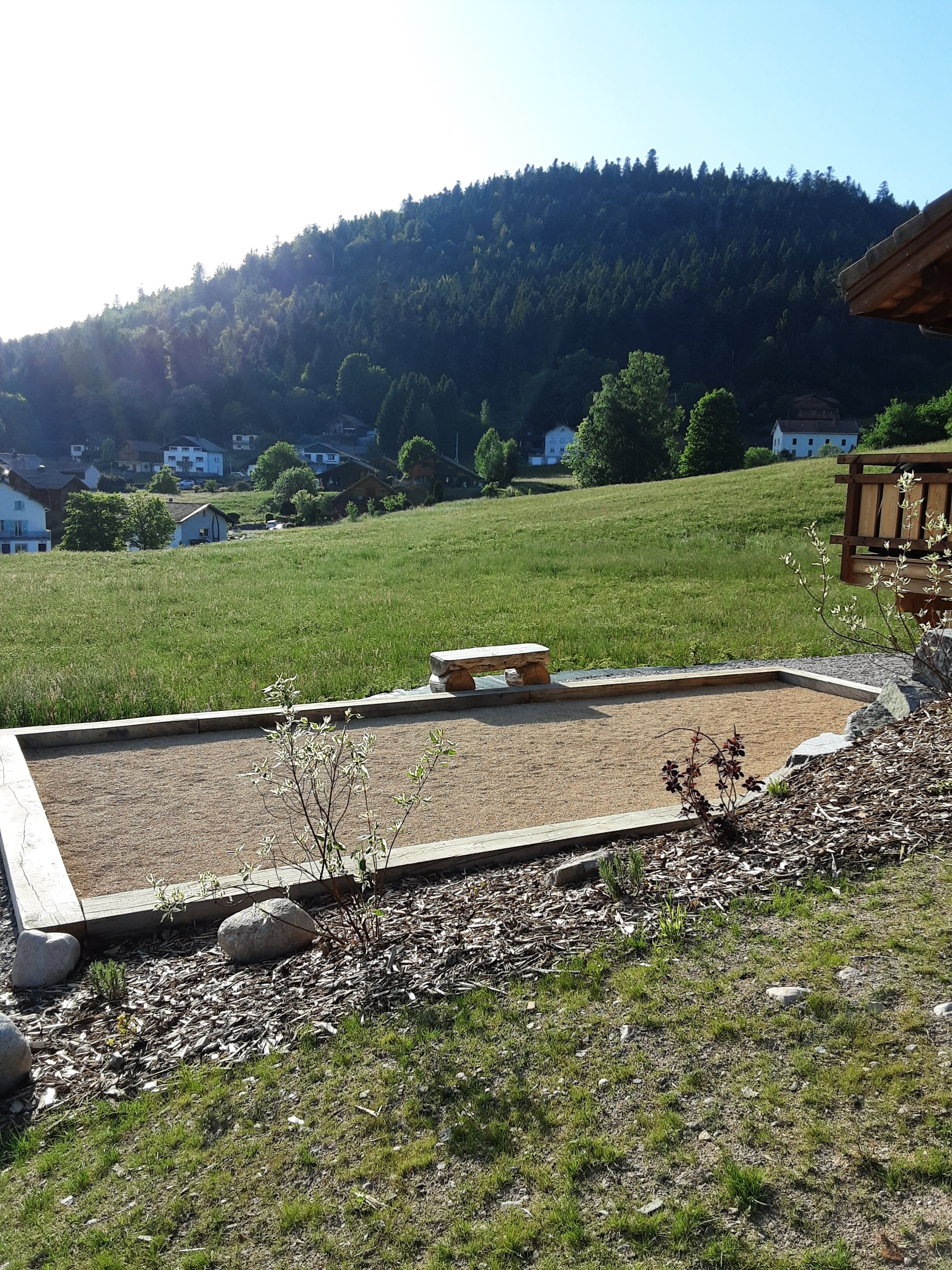 Un jardin avec un banc en bois, un parterre surélevé et une vue sur des maisons et une colline boisée en arrière-plan.