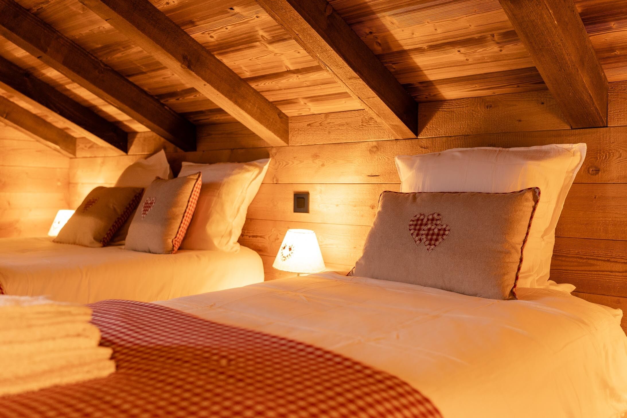 A cozy attic bedroom with wooden beams, two beds with white linens, and a small bedside lamp.