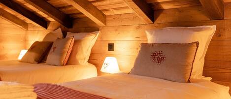 A cozy attic bedroom with wooden beams, two beds with white linens, and a small bedside lamp.