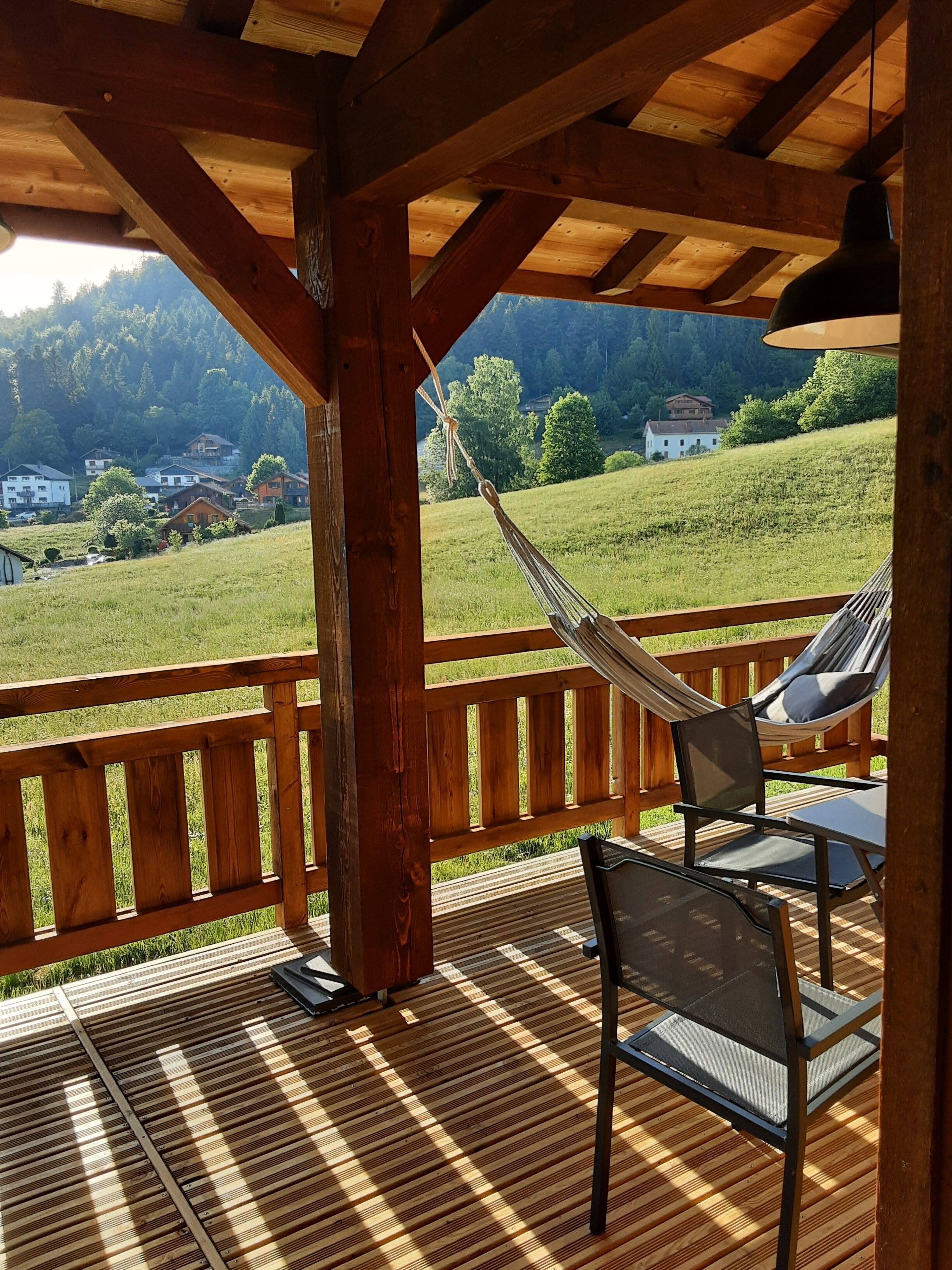 A wooden deck with a hammock and chairs, overlooking a green landscape with houses.