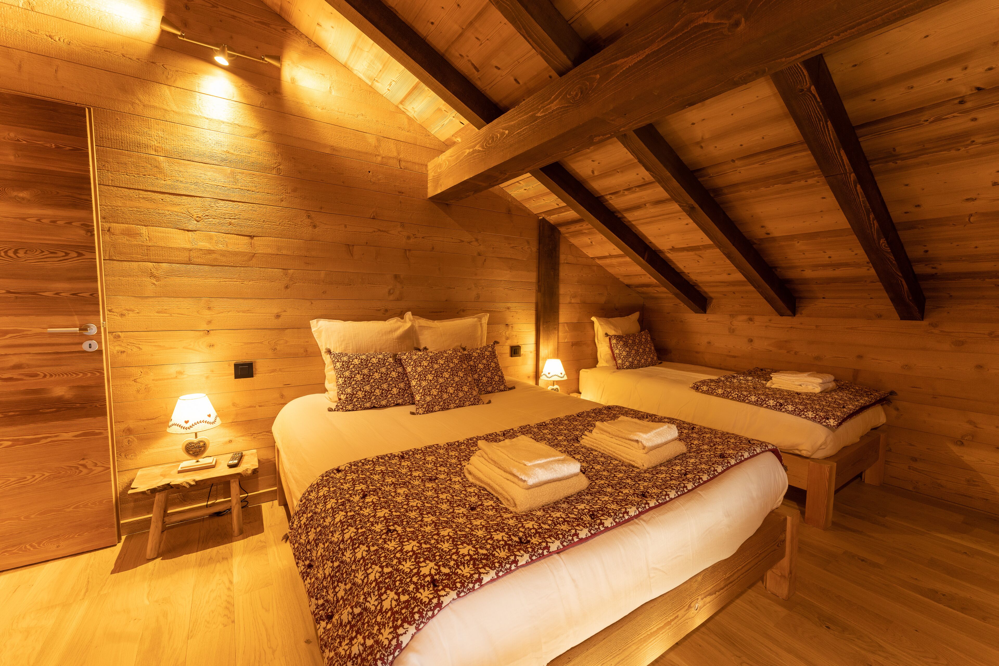 A wooden cabin bedroom with two beds, each with a patterned bedspread and pillows, a nightstand with a lamp, and a wooden ceiling with exposed beams.