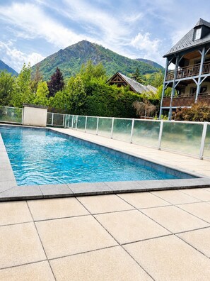 Una piscina con bordeado de baldosas, rodeada por una cerca blanca, con una montaña y un edificio al fondo.