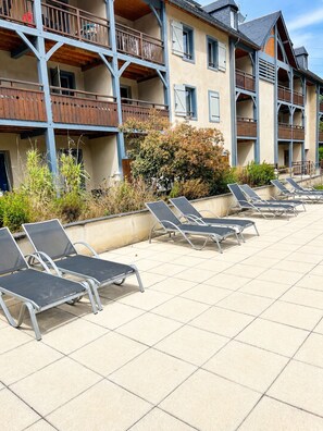 A building with multiple balconies, a tiled patio area with sun loungers, and a variety of plants.