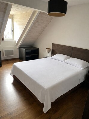 Un dormitorio con techo inclinado, una cama con ropa de cama blanca, una mesita de noche, una lámpara y una ventana.