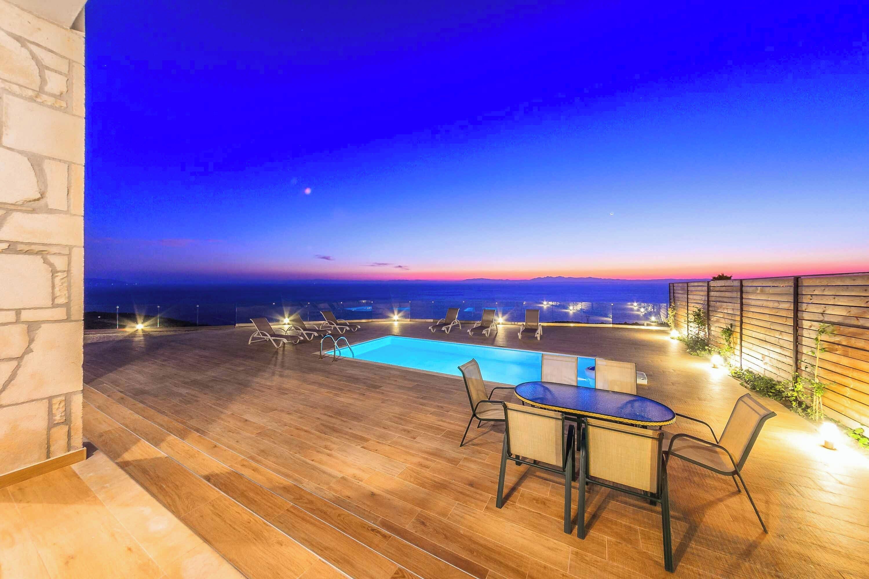 Una terraza de madera con piscina, mobiliario exterior y vista al horizonte al anochecer.