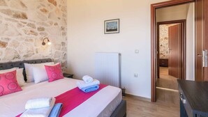 Un dormitori amb paret de pedra, un llit amb almoades, un radiador i una porta de fusta que condueix a una altra habitació.