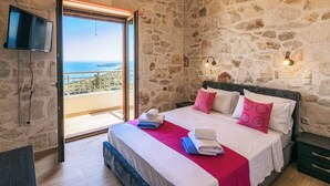 Un dormitori amb una paret de pedra, un llit amb roba de llit blanca i almoines roses, una manta vermella i vista al mar a través d'una porta de vidre.