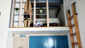 Une pièce compacte avec un espace en mezzanine décoré d’une planche de surf, comprenant une échelle et une étagère avec une voiture et des plantes.