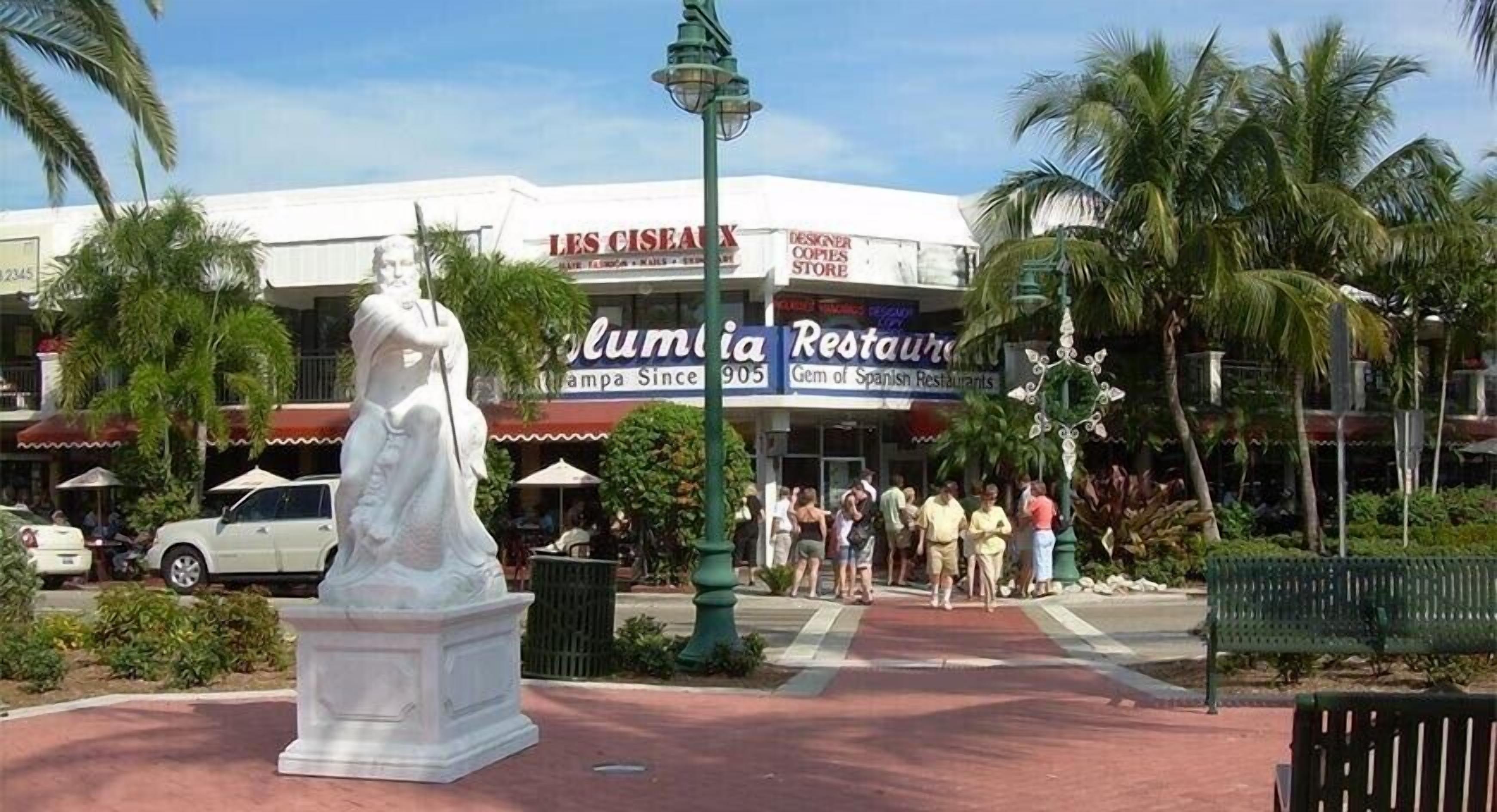 A statue in a plaza with palm trees and a restaurant in the background.