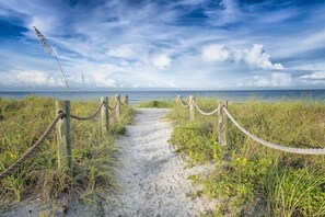 A sandy pathway leading to the beach, surrounded by grass and wooden posts with ropes.