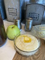 A bowl of oatmeal with a pat of butter on top, an apple, and two salt shakers labeled "Pepper" and "Flour" on a wooden surface.