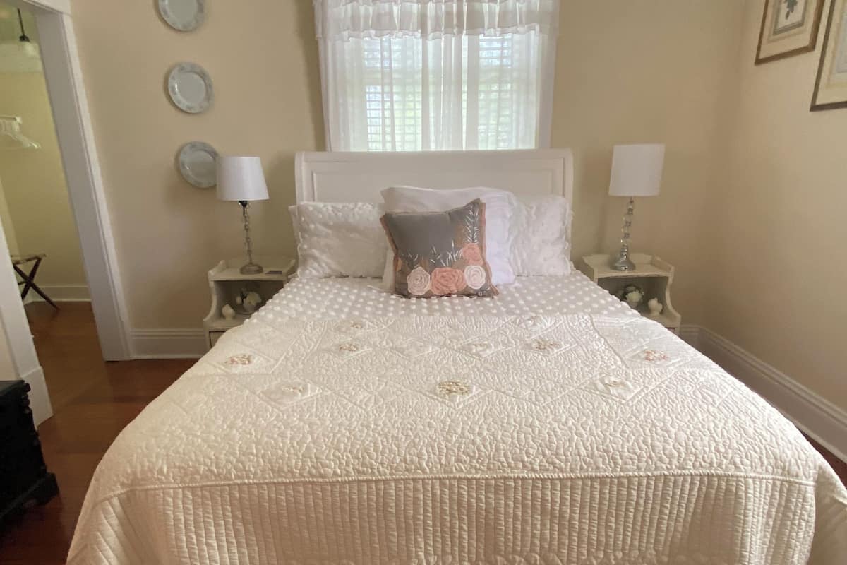 A neatly made bed with a white quilt, two bedside lamps, and a window with sheer curtains.