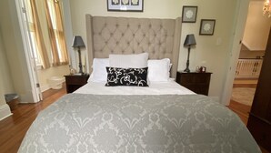 Un dormitorio con una cama grande, cabecera capitonada y colcha estampada. Hay dos mesitas de noche con lámparas y un cuadro enmarcado sobre la cama.