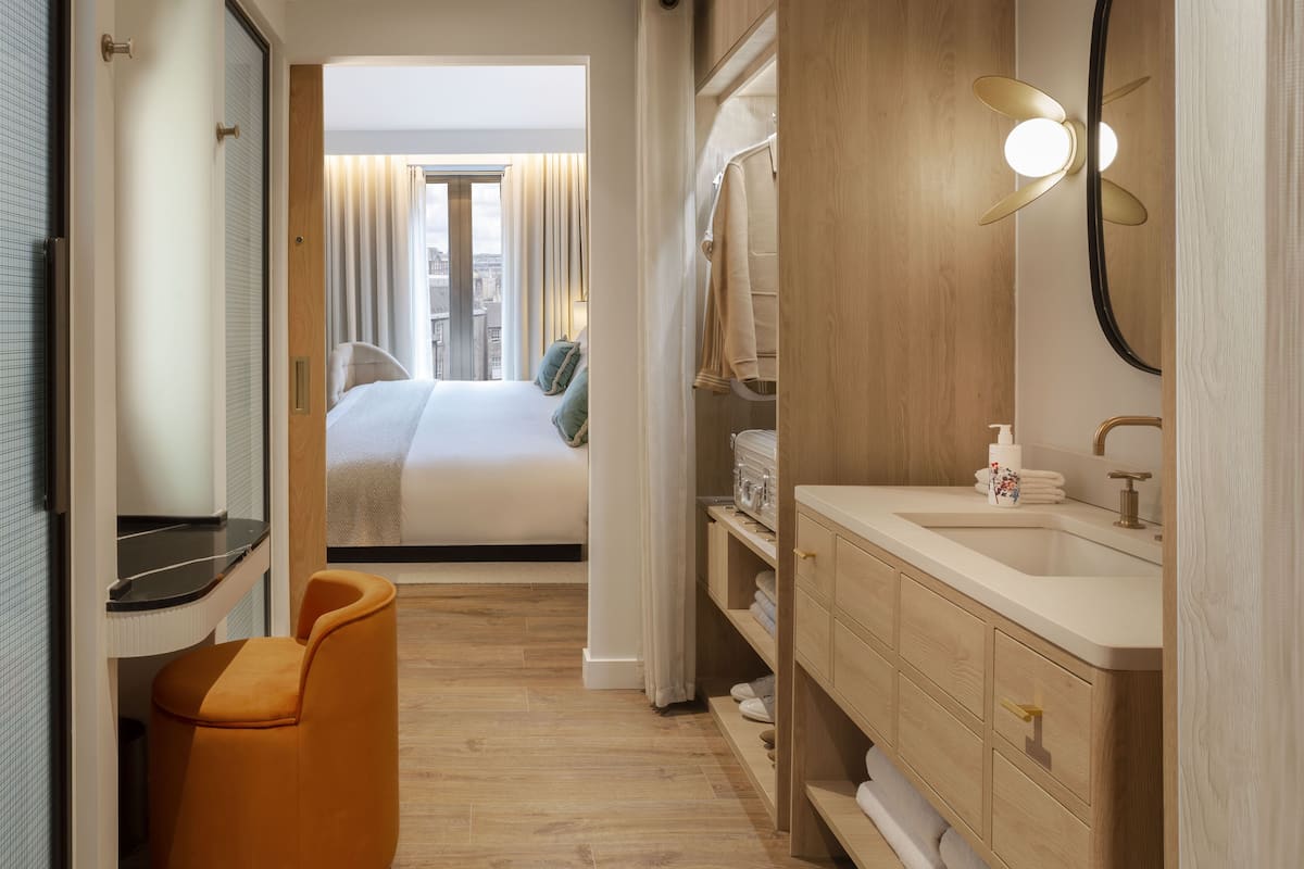 A hotel room with a bed, a bathroom with a sink and mirror, and a view of the city through the window.