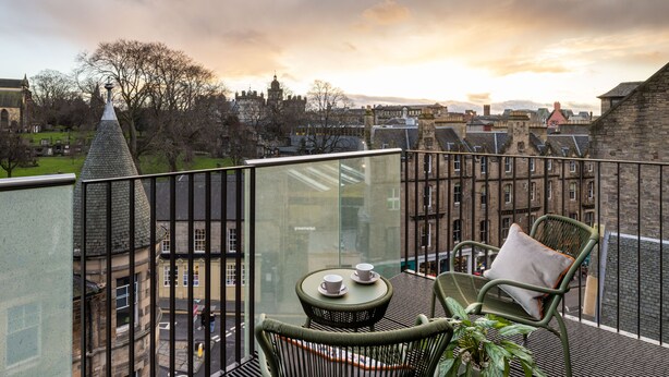 A balcony with a glass railing, two wicker chairs, a small round table with two cups, and a view of historic buildings and a church.