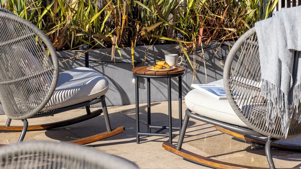 A rooftop terrace with wicker chairs, a small table with a cup and snacks, and a view of a castle-like building in the background.