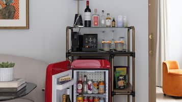 Un espacio habitable compacto con kitchenette, un refrigerador, una mesita con una planta y una puerta que da a otra habitación.