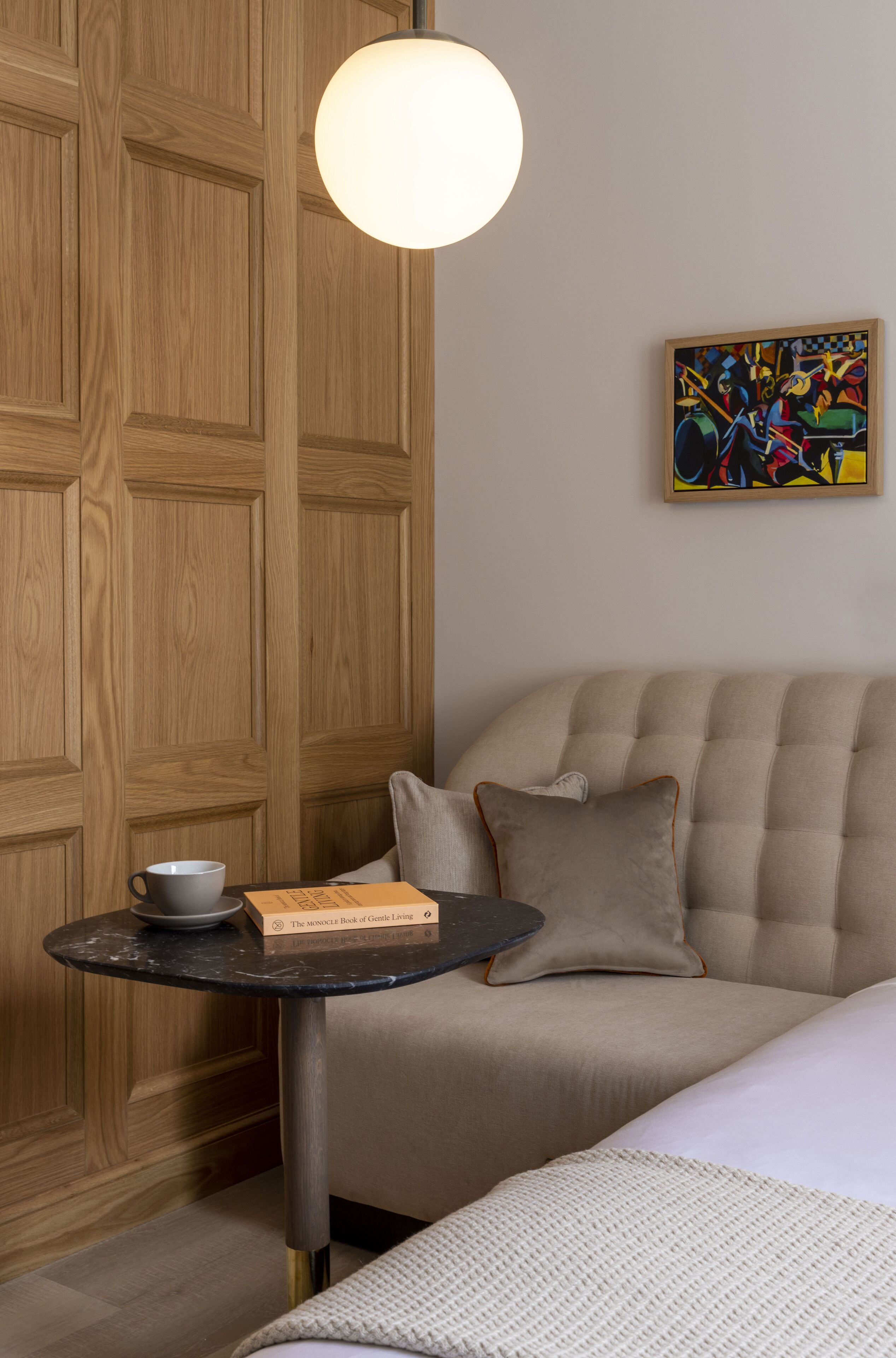 A cozy room with a wooden wardrobe, a round table with a book and a cup, a sofa with cushions, and a colorful abstract painting on the wall.