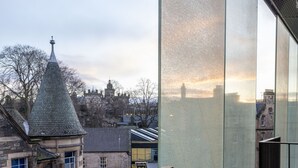 A modern building with large glass windows, a metal railing, and a view of a historic town with a tower.