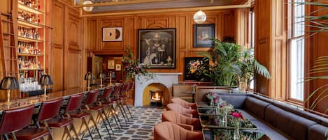 A bar area with red chairs and a long counter, a fireplace with a painting above it, and a seating area with a sofa and tables.