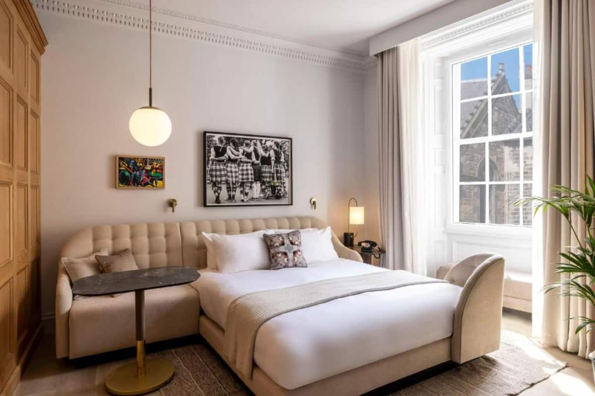 A hotel room with a large bed, a sofa, a small table, a lamp, and a window with curtains.