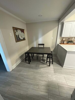 A dining area with a table and chairs, a painting on the wall, and a kitchen area with cabinets and a countertop.