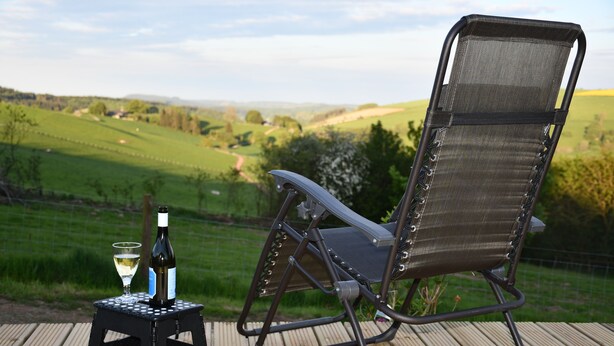 Ein Stuhl mit einer Weinflasche und einem Glas auf einer Holzterrasse mit Blick auf eine grüne Landschaft.