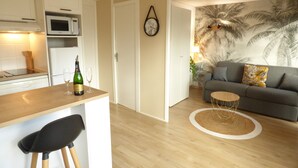 Una cocina moderna con microondas y una botella de vino en la encimera. Hay una zona de comedor con una silla negra y una mesa redonda de centro. La sala de estar tiene un sofá gris y un mural de una palmera en la pared.