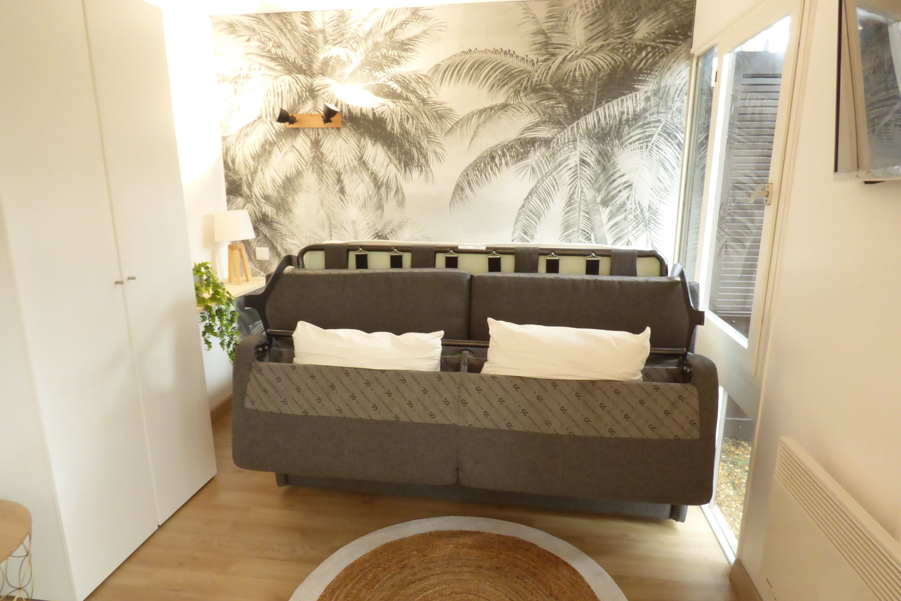 Un salon moderne avec un canapé gris, un plaid à motifs et une grande fenêtre donnant sur des palmiers.