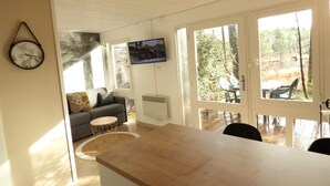 Sala de estar con sofá, mesa de centro y televisor. Amplias ventanas y vista al exterior.