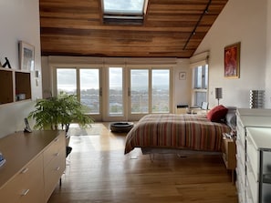 A bedroom with a large bed, a desk, a chair, a plant, and a view of the city through large windows. - Spacious home with 3 decks of spectacular views overlooking Provincetown Harbor (Provincetown)