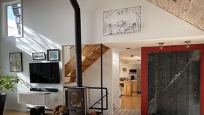 Ett modernt vardagsrum med en vedeldad kamin, en trappa och ett kök i bakgrunden.