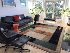 A modern living room with a black leather sofa, a red ottoman, a black office chair, and a colorful area rug. There are two large potted plants and a small table. - Spacious home with 3 decks of spectacular views overlooking Provincetown Harbor (Provincetown)