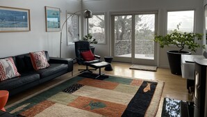 Ett rymligt vardagsrum med en svart lädersoffa, ett stort fönster med utsikt över träd, en svart stol med en röd kudde och en krukväxt.