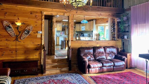 A cozy wooden cabin interior with a leather sofa, a dining area, and a kitchen visible through an open doorway. - Spacious Adirondack House in Otter Lake (Forestport)