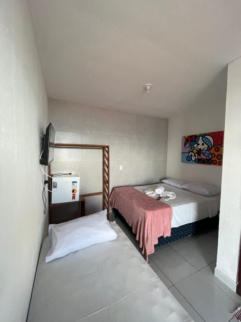 Habitació d'hotel petita i senzilla amb un llit, una taula de nit, una televisió i un frigorífic.