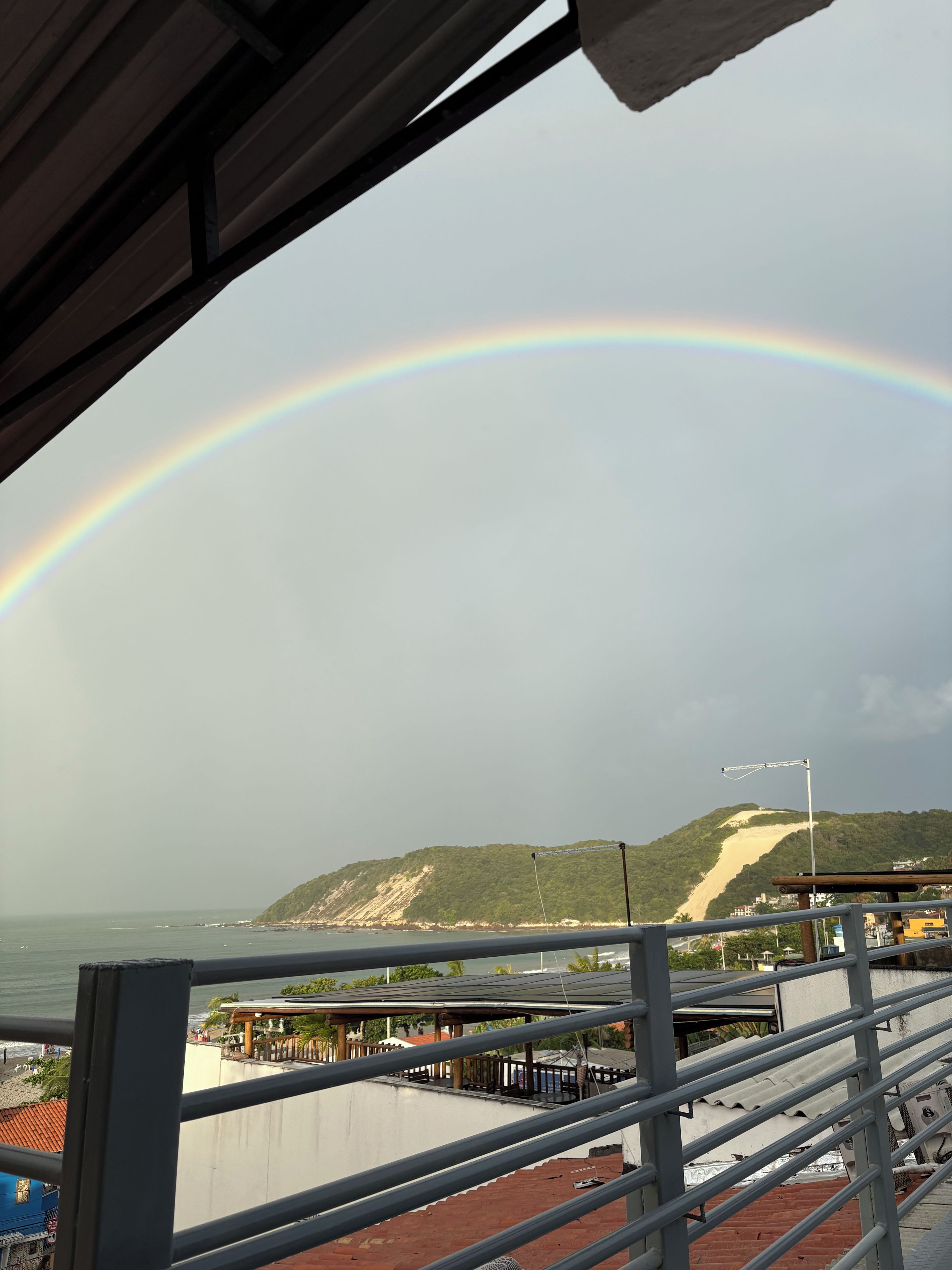 Vista di una spiaggia con un arcobaleno, una struttura in legno e una ringhiera metallica.