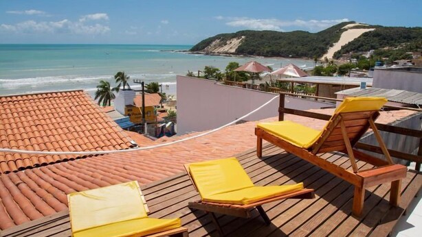 A wooden deck with yellow lounge chairs overlooking a beach with a clear view of the ocean and a hill in the distance. - Hotel Pousada Encanto da Praia  (Natal)