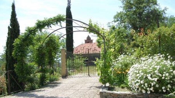 Un sentiero sterrato conduce a un edificio con tetto in tegole rosse, immerso nel verde e sormontato da un arco decorativo.