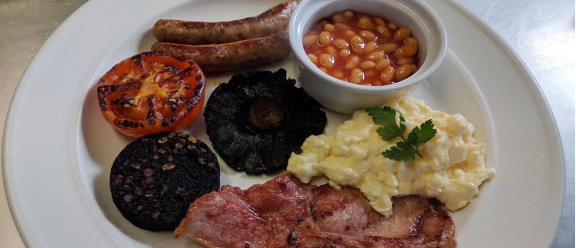 A plate with grilled tomato, black pudding, sausages, baked beans, scrambled eggs, and bacon.