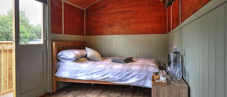 A single bed with a wooden headboard, a bedside table with a water bottle and glasses, and a view of the outdoors through a large window.