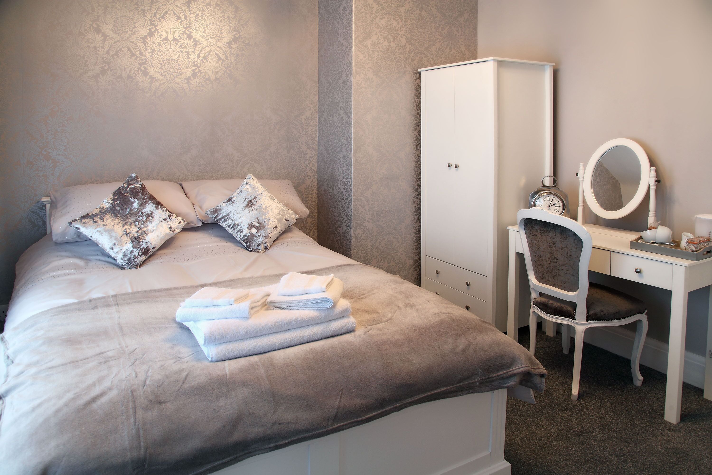 A neatly made bed with pillows, a white wardrobe, a chair, and a desk with a mirror and clock.