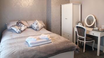 Een opgemaakt bed met kussens, een witte kledingkast, een stoel en een bureau met spiegel en klok.