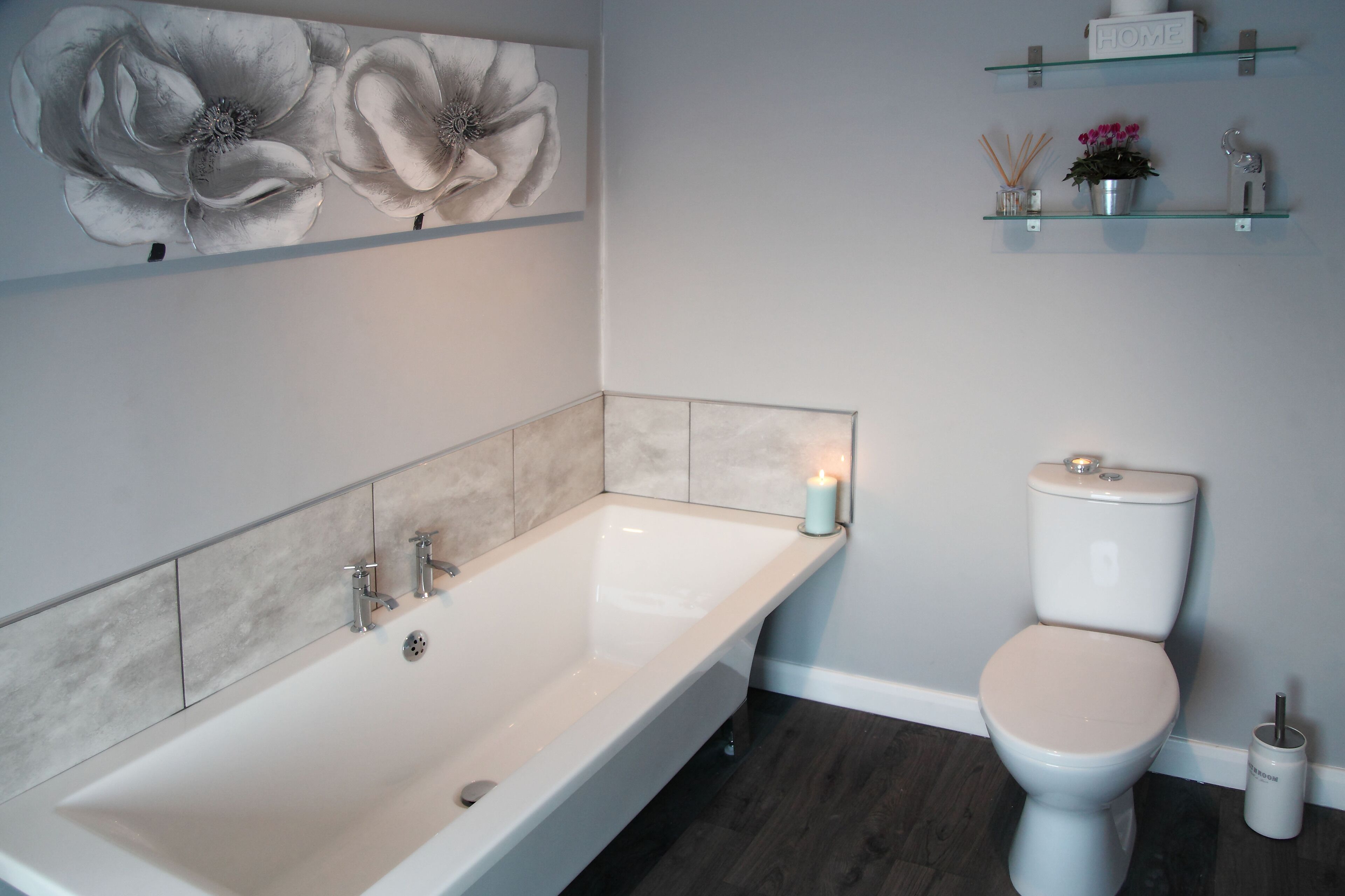 A modern bathroom with a bathtub, toilet, and decorative elements including a flower painting and a shelf with toiletries.