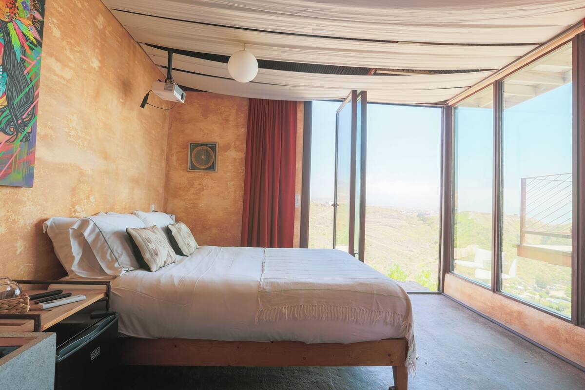 A bedroom with a large bed, a desk with a lamp, and a view of the landscape through large windows.