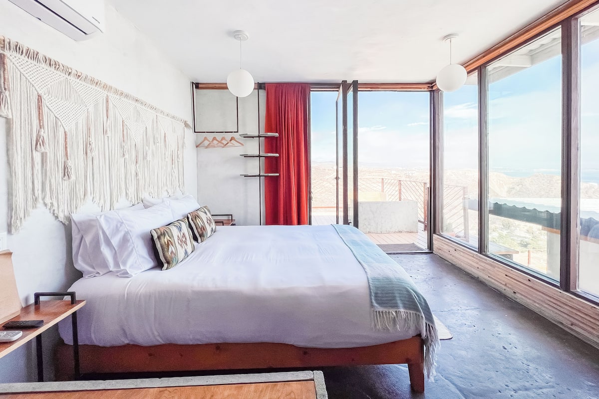 A modern bedroom with a large bed, a macrame wall hanging, a red curtain, and a balcony with a city view.